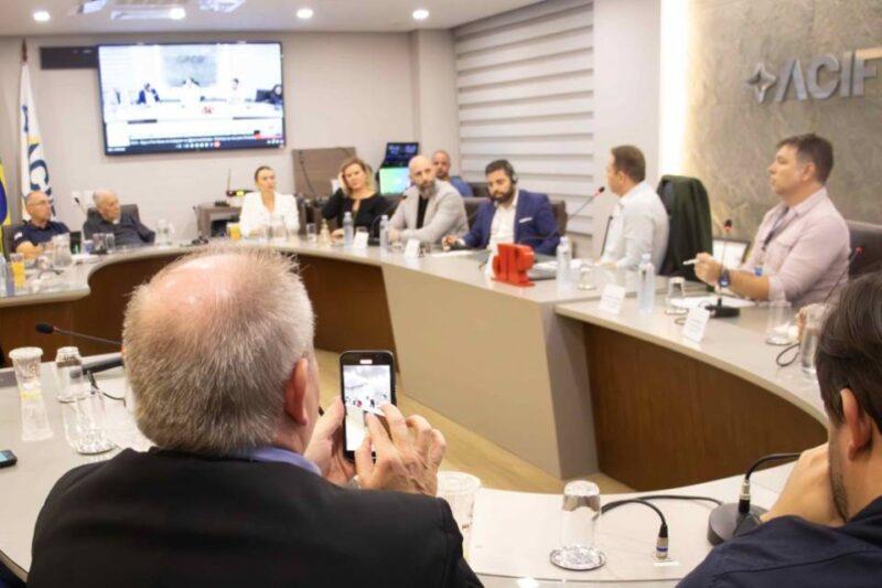 Sala de reunião moderna com uma mesa em formato de U, onde vários participantes estão sentados, discutindo em um painel ou conferência. Ao fundo, uma tela grande exibe imagens que parecem ser da própria reunião. À esquerda, uma pessoa de cabelos grisalhos usa um celular para fotografar ou gravar o evento. O ambiente é formal, com microfones, copos de água e documentos distribuídos sobre a mesa. O logotipo da ACIF (Associação Comercial e Industrial de Florianópolis) é visível na parede ao fundo, indicando que a reunião está relacionada à organização.