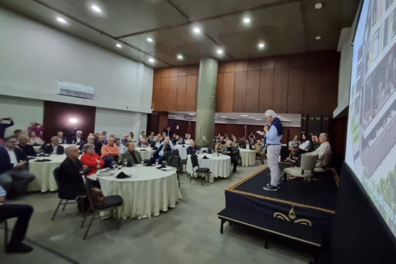 Sala de conferências com diversas mesas redondas onde estão sentadas várias pessoas, principalmente homens e mulheres de meia-idade. No canto direito, um homem mais velho está em pé, em um palco pequeno, falando ao microfone enquanto apresenta um slide projetado na parede. A plateia parece atenta, e há uma atmosfera formal e organizada, típica de eventos corporativos ou institucionais. O ambiente é bem iluminado, com iluminação artificial de teto e decoração sóbria.