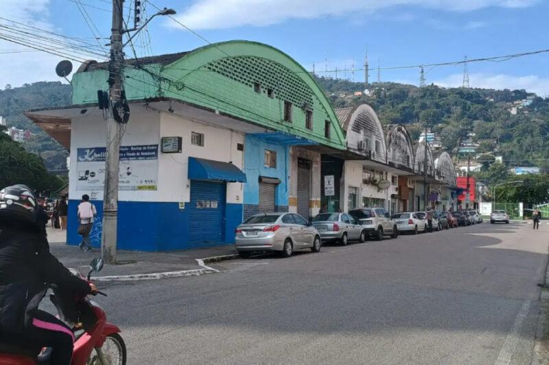 "Vista de uma rua urbana com uma fileira de galpões antigos de arquitetura industrial ao longo do lado direito. Os galpões têm fachadas coloridas, principalmente em tons de verde e azul, com telhados curvos e janelas pequenas. Alguns carros estão estacionados na calçada ao lado dos galpões. No canto esquerdo da imagem, uma pessoa em uma motocicleta é vista passando pela rua. Ao fundo, é possível ver uma colina coberta de vegetação com várias antenas de comunicação no topo, sob um céu parcialmente nublado."