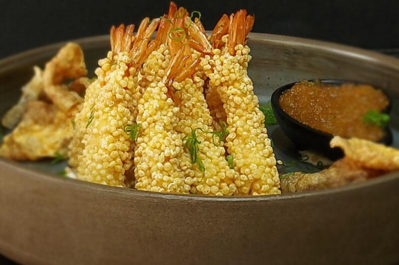 "Prato gourmet com camarões empanados em crosta de quinoa, dispostos verticalmente em um prato rústico. Ao lado, pedaços de peixe crocante e um molho de acompanhamento em um recipiente pequeno. A apresentação do prato é sofisticada, com detalhes verdes de cebolinha salpicados sobre os camarões."