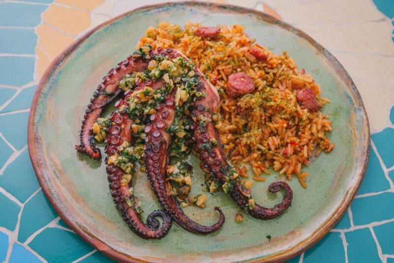 "Prato de polvo grelhado coberto com molho verde de ervas e vegetais, acompanhado por arroz com pedaços de linguiça. A comida está disposta em um prato rústico verde sobre uma mesa colorida com mosaico, destacando a apresentação vibrante e apetitosa do prato."