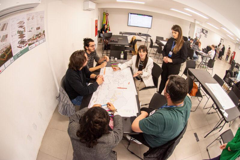 Um grupo de seis pessoas está reunido em torno de uma mesa retangular em uma sala de reunião. Eles estão discutindo e desenhando em um mapa grande que cobre a mesa. Uma mulher de pé, com crachá de identificação, observa a discussão. A sala é clara e moderna, com paredes brancas e outros grupos trabalhando em mesas ao fundo. No fundo da sala, há bandeiras do Brasil e de Santa Catarina, além de um monitor de TV exibindo informações.