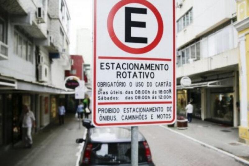 A imagem mostra uma placa de trânsito de Estacionamento Rotativo em uma rua urbana. A placa é predominantemente branca com bordas e texto em preto, exceto pelo símbolo de "E" de estacionamento dentro de um círculo vermelho no topo. Abaixo do símbolo, o texto informa que é obrigatório o uso do cartão de estacionamento, indicando os horários de funcionamento: de segunda a sexta-feira, das 08h00 às 18h00, e aos sábados, das 08h00 às 12h00. No rodapé da placa, há uma restrição informando que é proibido o estacionamento de ônibus, caminhões e motos. Ao fundo, é possível ver uma rua comercial com pessoas caminhando e lojas dos dois lados.