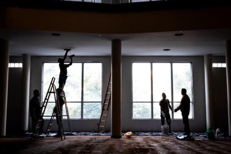 "Quatro trabalhadores são vistos realizando atividades de manutenção no interior de um edifício. Um deles está em uma escada, mexendo no teto, enquanto outros três estão no chão, conversando e organizando ferramentas. A iluminação natural entra pelas grandes janelas ao fundo, criando um contraste com o ambiente interno mais escuro. Escadas e ferramentas estão espalhadas pelo local."