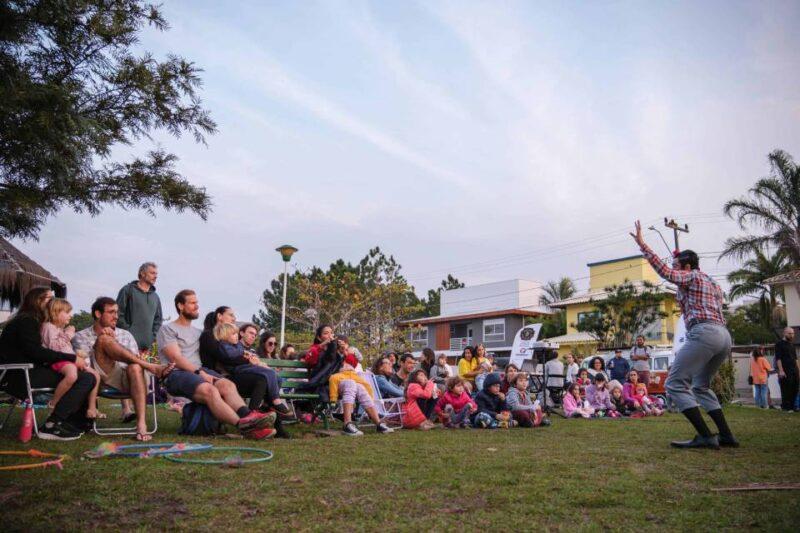 Um grupo de pessoas, incluindo adultos e crianças, está sentado em cadeiras e no chão em um gramado, assistindo a uma performance ao ar livre. À direita da imagem, um artista vestindo camisa xadrez e calças claras gesticula energicamente durante a apresentação. Ao fundo, casas coloridas e vegetação típica de um bairro residencial. A atmosfera é tranquila, com o público atento e envolvido na apresentação, que parece ocorrer ao entardecer.