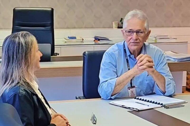 Duas pessoas estão sentadas em uma mesa de reunião em um ambiente corporativo. À direita, um homem idoso de cabelos grisalhos, usando óculos e camisa azul clara, está com as mãos cruzadas sobre a mesa, olhando atentamente para a pessoa à sua esquerda. À esquerda, uma mulher de cabelos grisalhos e longos, vestindo uma jaqueta escura, está de perfil, sorrindo enquanto conversa. Na mesa, há documentos e pastas organizadas. O ambiente sugere uma discussão ou reunião formal em um escritório.