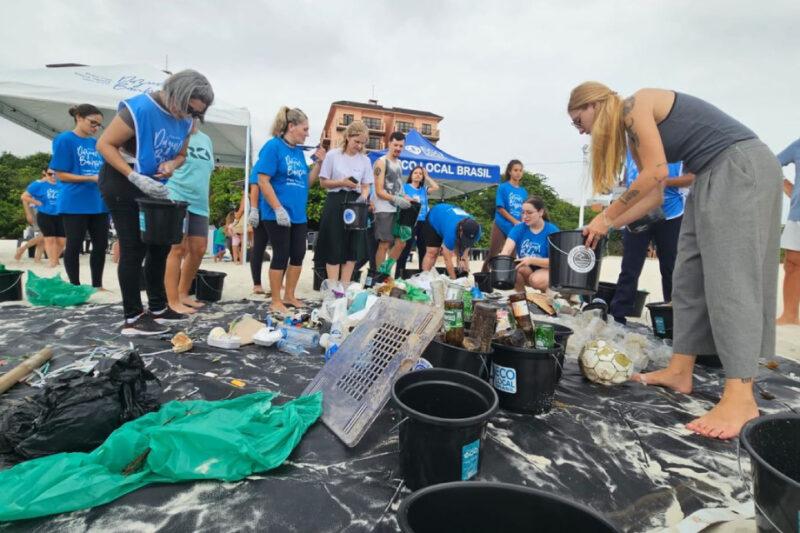 "Voluntários participam de uma ação de limpeza de praia, recolhendo e separando lixo acumulado na areia. Eles estão vestidos com camisetas azuis com o logo da campanha e utilizam baldes pretos para coletar os resíduos. Há diversos itens espalhados no chão, como garrafas plásticas, latas e outros detritos. Ao fundo, é possível ver uma tenda branca com a marca de um patrocinador e algumas construções ao longe. O ambiente é de colaboração e conscientização ambiental."