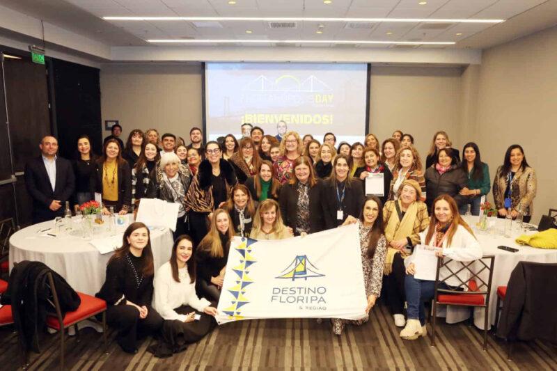 "Foto de um grande grupo de pessoas reunidas em uma sala de conferências, posando para a câmera. A maioria dos participantes são mulheres, e todas estão sorrindo. Na frente do grupo, algumas pessoas seguram uma faixa com o logo e o nome 'Destino Floripa & Região'. Ao fundo, há um projetor exibindo as palavras 'Florianópolis Day' e 'Bienvenidos!' em um slide. A sala é moderna, com cadeiras e mesas redondas, decoradas com arranjos de flores e copos. O ambiente é acolhedor e profissional, sugerindo um evento ou encontro corporativo."