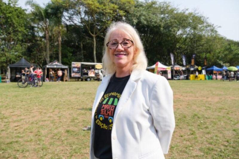 "Uma mulher idosa com cabelos brancos e óculos está sorrindo em um parque durante o dia. Ela veste um blazer branco sobre uma camiseta preta que tem o logotipo colorido de um evento chamado 'Mercado Aberto - Economia Criativa'. Ao fundo, há barracas de eventos e pessoas andando de bicicleta ou conversando em um ambiente arborizado."