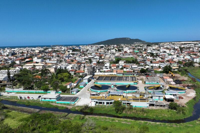 Vista aérea de uma estação de tratamento de esgoto localizada no bairro Ingleses, em Florianópolis, SC. A estação ocupa a parte inferior da imagem e é composta por diversas estruturas circulares e retangulares, cercadas por prédios e vegetação. Ao fundo, vê-se o bairro residencial com muitas casas de telhados alaranjados e edifícios baixos. Mais ao fundo, destacam-se as montanhas e o mar, sob um céu azul claro. Um pequeno rio serpenteia pelo lado direito da imagem, atravessando áreas verdes até desaparecer no horizonte.