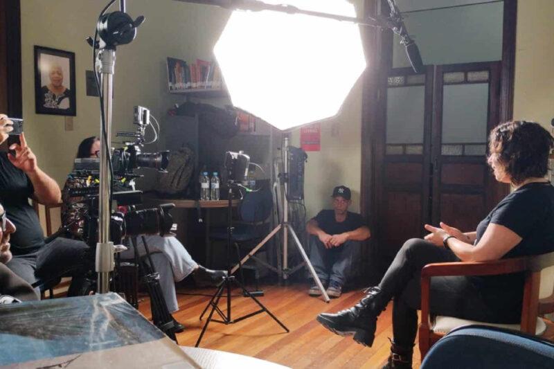 A imagem mostra uma cena de gravação em um ambiente interno. Uma mulher, sentada à direita da imagem, está sendo entrevistada ou filmada. Ela está de perfil e veste roupas pretas. Ao redor dela, há equipamentos de filmagem como câmeras e tripés, com uma luz de estúdio grande e brilhante direcionada para ela, posicionada no centro da imagem. À esquerda, membros da equipe técnica ajustam os equipamentos enquanto outro profissional está sentado no chão, encostado na parede, observando a cena. No fundo, há uma porta de madeira escura e algumas estantes com livros, além de uma foto emoldurada na parede.