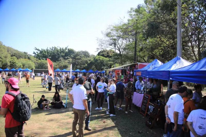 "Foto de um evento ao ar livre com várias barracas azuis dispostas em fileiras, onde as pessoas estão interagindo e comprando produtos. Muitas pessoas estão presentes, algumas caminhando, outras conversando e algumas sentadas na grama. Há árvores e vegetação ao fundo, sugerindo um ambiente de parque ou área verde. O céu está claro e ensolarado, indicando que é um dia agradável."