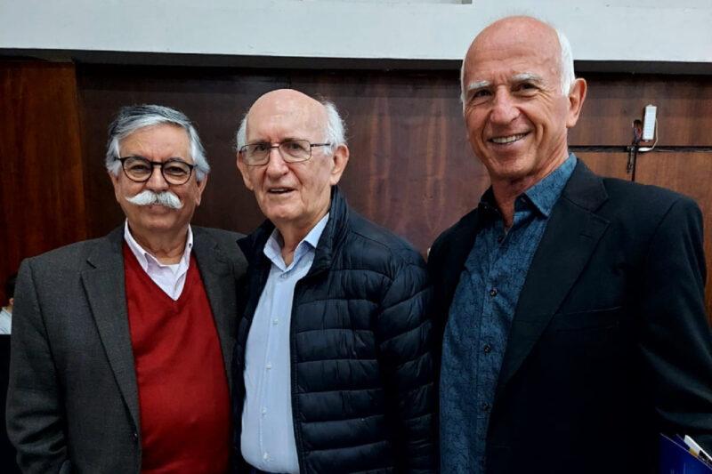 Três homens sorridentes posam juntos para a foto em um ambiente interno. O homem à esquerda tem cabelos grisalhos, usa óculos, um bigode marcante e veste um suéter vermelho com blazer cinza. O homem no centro, de cabelos brancos, usa óculos e está com uma jaqueta preta sobre uma camisa azul clara. O homem à direita é calvo e veste um blazer preto com camisa azul escura. O fundo é neutro, com painéis de madeira, indicando um ambiente formal, possivelmente em um evento ou reunião.