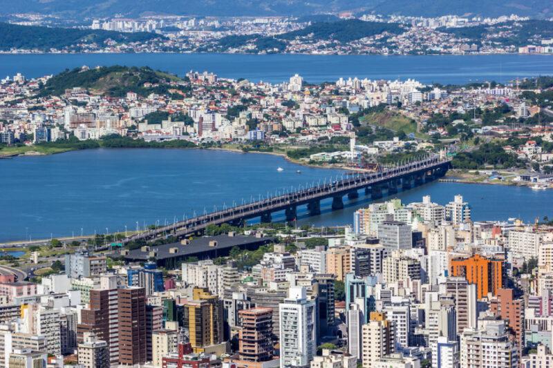 "Vista aérea de Florianópolis, mostrando a Ponte Colombo Salles atravessando a Baía Norte e conectando a ilha ao continente. Em primeiro plano, há vários prédios altos no centro da cidade, enquanto ao fundo é possível ver a parte continental da cidade com morros verdes e áreas residenciais. A imagem captura a geografia única da região, com suas águas azuis e a urbanização densa tanto na ilha quanto no continente."