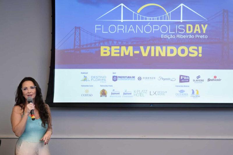 "Uma mulher de cabelos longos e castanhos, vestindo uma blusa verde e calça bege, segura um microfone enquanto fala em frente a uma tela de projeção. A tela exibe o título 'Florianópolis Day - Edição Ribeirão Preto' com uma imagem da Ponte Hercílio Luz ao fundo e a palavra 'BEM-VINDOS!' em destaque. A parte inferior da tela contém logos de diversos patrocinadores e apoiadores do evento."