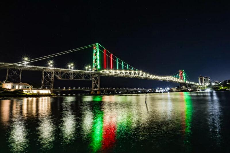 Foto noturna da Ponte Hercílio Luz, em Florianópolis, iluminada com luzes nas cores verde e vermelha. As luzes destacam a estrutura da ponte contra o céu escuro, refletindo na água abaixo, criando um efeito visual vibrante e colorido. No lado esquerdo, é possível ver parte da orla com algumas construções iluminadas, e no fundo, há prédios da cidade. A imagem transmite a beleza e a grandiosidade da ponte histórica sob uma iluminação especial.