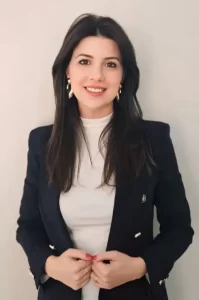 Retrato de uma mulher jovem, com cabelos castanhos longos e soltos, usando um blazer preto sobre uma blusa branca de gola alta. Ela está sorrindo, com as mãos tocando o blazer na altura da cintura. Ao fundo, uma parede de cor neutra