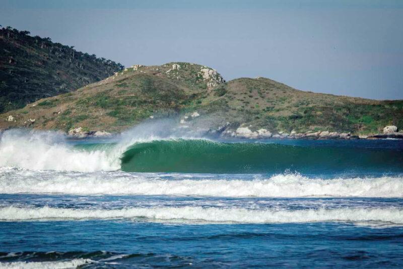 "Uma bela paisagem da praia de Moçambique, em Florianópolis. A imagem captura uma onda verde cristalina quebrando na praia, com espuma branca ao redor. Ao fundo, há uma formação rochosa com vegetação esparsa, destacando a natureza preservada do local. O céu está claro, sugerindo um dia ensolarado."