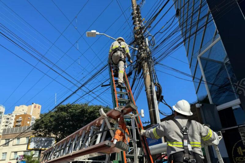 "Dois trabalhadores da Celesc, equipados com uniformes de segurança e capacetes, estão realizando manutenção em um poste de energia elétrica. Um dos trabalhadores está no topo de uma escada apoiada no poste, enquanto o outro o assiste a partir do chão. O poste está rodeado por uma densa rede de cabos, e o céu azul sem nuvens ao fundo destaca a cena. Ao redor, podem ser vistos prédios residenciais e comerciais."