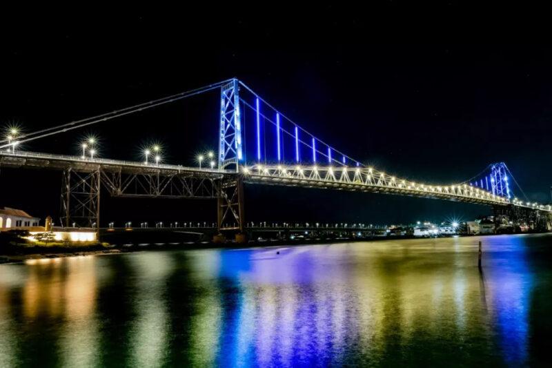 "Fotografia noturna da Ponte Hercílio Luz em Florianópolis, iluminada com luzes azuis e brancas. A estrutura metálica da ponte se reflete na água tranquila abaixo, criando um efeito de espelhamento colorido. O céu está escuro, destacando ainda mais as luzes da ponte."