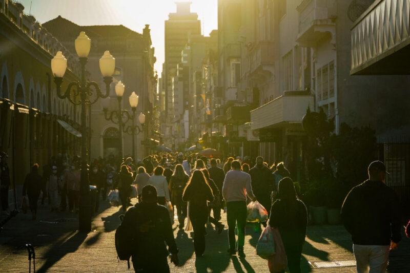 "Uma rua movimentada no centro de uma cidade ao entardecer, com muitas pessoas caminhando em direção à câmera. O sol baixo no horizonte cria silhuetas e longas sombras das pessoas e dos edifícios ao redor. As luzes dos postes de estilo antigo estão apagadas, e as fachadas dos prédios refletem a luz dourada do sol. A cena transmite uma sensação de movimentação urbana e de fim de tarde."