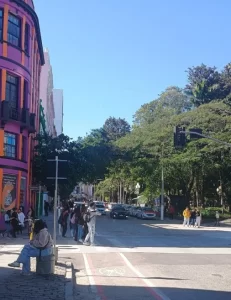 Imagem de uma movimentada rua em Florianópolis, com várias pessoas andando nas calçadas e atravessando a faixa de pedestres. À esquerda, um prédio colorido com tons vibrantes de rosa e laranja se destaca. No fundo, árvores verdes compõem o cenário, e carros estão parados no semáforo. O céu está claro e azul, indicando um dia ensolarado.