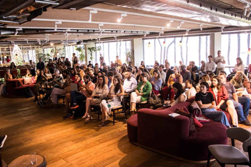 "Imagem de um auditório moderno e bem iluminado, com um grande grupo de pessoas sentadas, assistindo a uma apresentação. O público é diverso, com homens e mulheres de diferentes idades. O ambiente tem piso de madeira, sofás e cadeiras confortáveis, e é decorado com plantas e grandes janelas que permitem a entrada de luz natural. As pessoas parecem atentas, algumas tirando notas ou usando dispositivos eletrônicos."