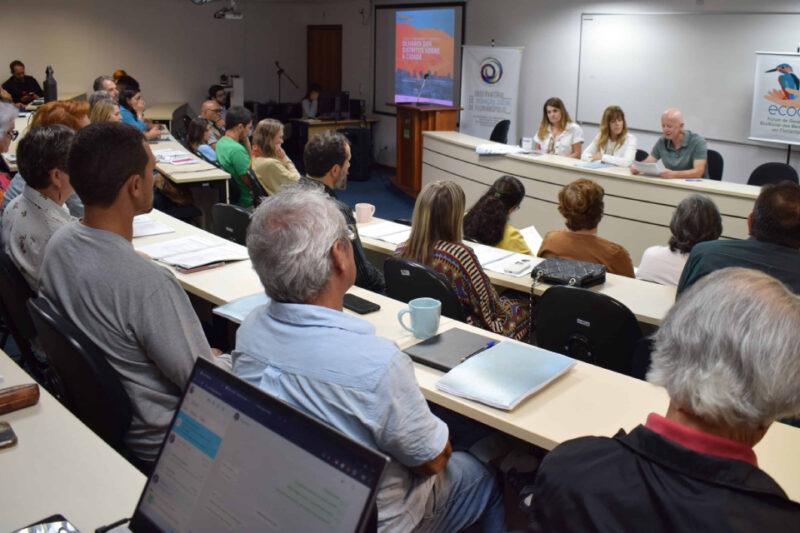 "Uma sala de conferências com várias pessoas sentadas em fileiras, prestando atenção a uma apresentação. Na frente, três palestrantes, duas mulheres e um homem, estão sentados em uma mesa, discutindo tópicos. Há um projetor exibindo uma apresentação na tela ao fundo com o título 'Olhares dos Visitantes sobre a Cidade'. Algumas pessoas na audiência estão fazendo anotações e há laptops abertos em algumas mesas. A atmosfera é de concentração e aprendizado."