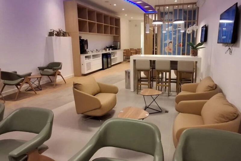 Lounge moderno com diversas poltronas e cadeiras estofadas em tons de bege e verde claro, dispostas ao redor de mesas pequenas. Ao fundo, há uma área de café com uma bancada alta, prateleiras abertas, e equipamentos como cafeteira e frigobar. A iluminação é composta por luminárias pendentes e luzes embutidas no teto