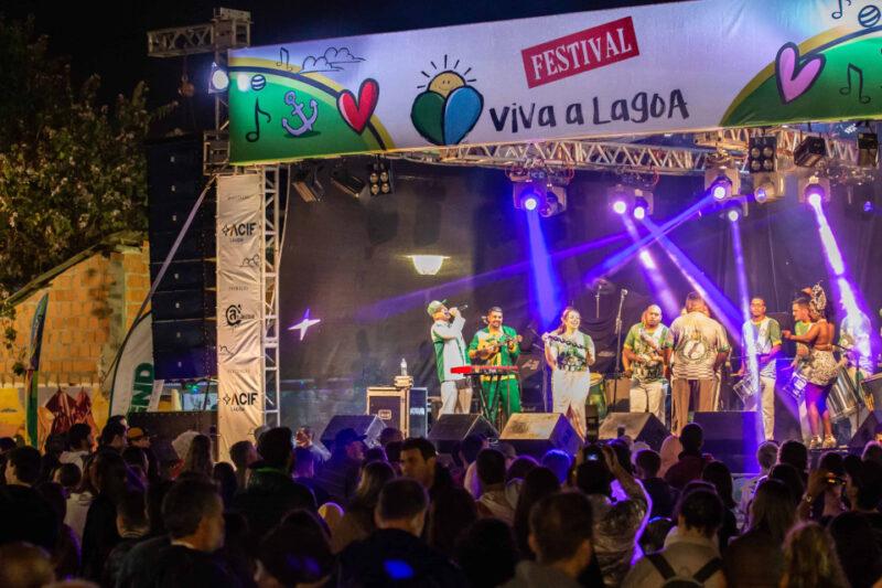 "Festival Viva a Lagoa com uma grande multidão assistindo a uma apresentação musical ao vivo à noite. O palco está iluminado com luzes coloridas e uma faixa decorativa no topo com o nome do evento. Os músicos, vestidos com trajes vibrantes, estão tocando instrumentos e cantando. O público se reúne em frente ao palco, criando um ambiente animado e festivo."