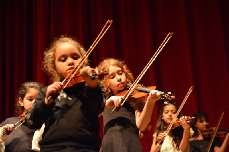 "Grupo de crianças tocando violino em uma apresentação. As crianças estão em foco no palco, com expressão concentrada enquanto manuseiam os instrumentos de cordas. Ao fundo, uma cortina vermelha cobre todo o cenário, destacando a performance musical."