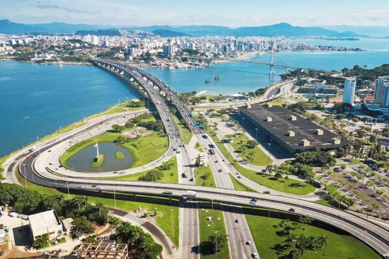 Vista aérea da cidade de Florianópolis, destacando a ponte Colombo Salles, que conecta a ilha ao continente. A imagem mostra as vias expressas bem definidas, com múltiplas faixas e fluxo de veículos, além de áreas verdes ao redor das estradas. À esquerda, há um pequeno lago com uma escultura no centro e árvores próximas. Ao fundo, é possível ver a cidade com muitos prédios e, à direita, a icônica ponte Hercílio Luz. O mar azul e as montanhas ao fundo completam a paisagem, sob um céu claro.