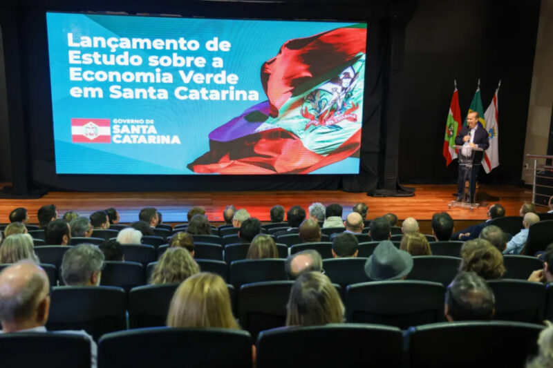 Fotografia de uma conferência com várias pessoas sentadas assistindo à apresentação de um estudo sobre Economia Verde em Santa Catarina. No palco, um palestrante está de pé ao lado de um púlpito transparente, com bandeiras de Santa Catarina e do Brasil ao fundo. Ao lado, uma grande tela exibe a mensagem: 'Lançamento de Estudo sobre a Economia Verde em Santa Catarina', junto ao logotipo do Governo de Santa Catarina e uma imagem da bandeira estadual.