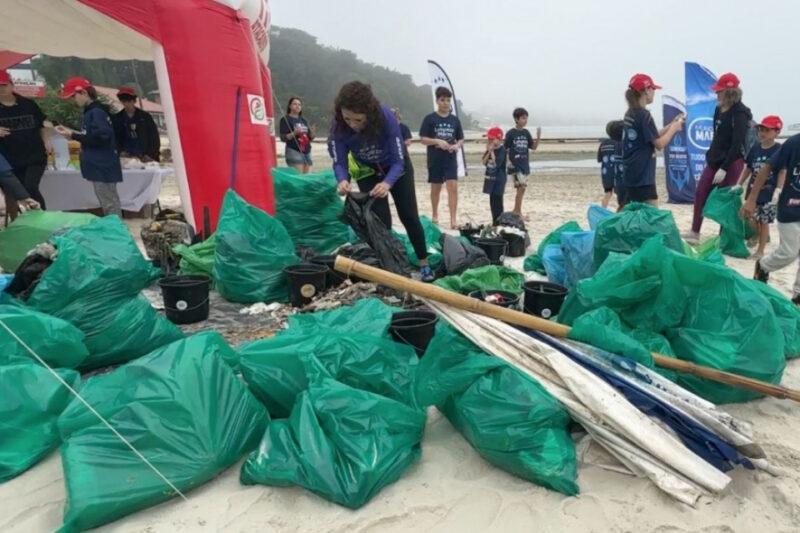 "Um grupo de pessoas, incluindo adultos e crianças, participa de uma ação de limpeza de praia. No centro da imagem, uma mulher de cabelo cacheado, vestindo uma camiseta azul e calça escura, está amarrando um saco verde cheio de lixo. Ao redor, há vários sacos verdes cheios de resíduos coletados e baldes pretos. Algumas pessoas estão usando bonés vermelhos e camisetas azuis com inscrições. Ao fundo, é possível ver um arco inflável vermelho e branco, possivelmente uma tenda ou entrada de um evento, além de banners azuis ao longe. O céu está nublado e a praia parece estar coberta por uma névoa leve, com o mar ao fundo."