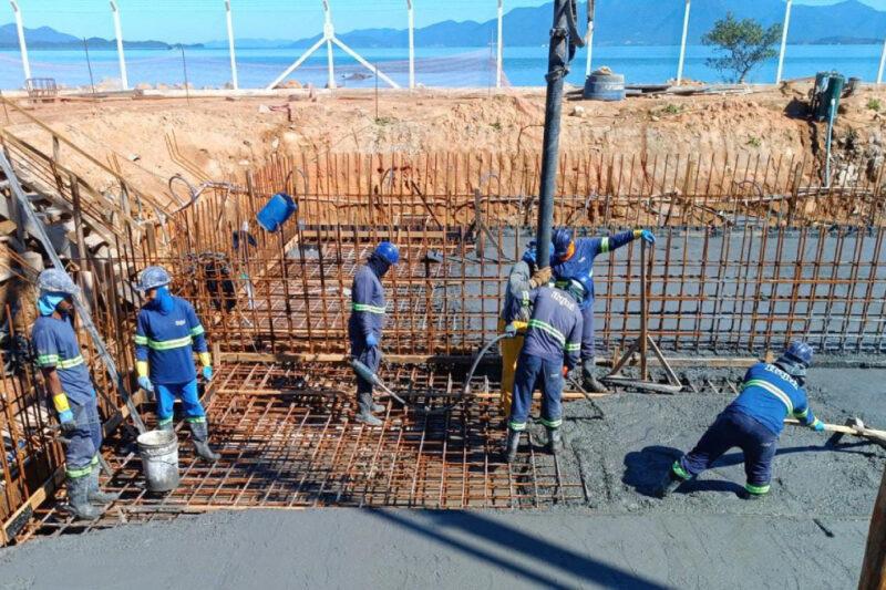 "Cinco trabalhadores da construção civil, usando uniformes azuis, capacetes e equipamentos de segurança, estão envolvidos no trabalho de concretagem de uma estrutura metálica em uma obra ao ar livre. Eles estão derramando e espalhando concreto fresco sobre a malha de aço, que será usada para reforçar a estrutura. Ao fundo, há uma cerca branca e uma vista de um corpo d'água cercado por montanhas, indicando que a obra está localizada perto de uma área costeira ou ilha."