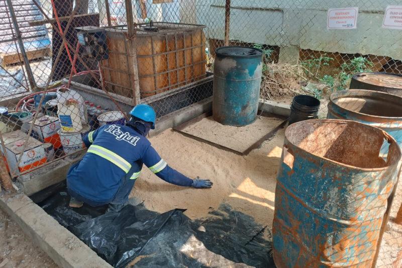 Trabalhador vestindo uniforme azul e capacete de proteção realiza a limpeza de um local com resíduos contaminados. Ele espalha um material de absorção no chão. O ambiente é cercado por tambores metálicos enferrujados e tanques de armazenamento. No fundo, há sinalizações indicando 'Resíduos Contaminados'.