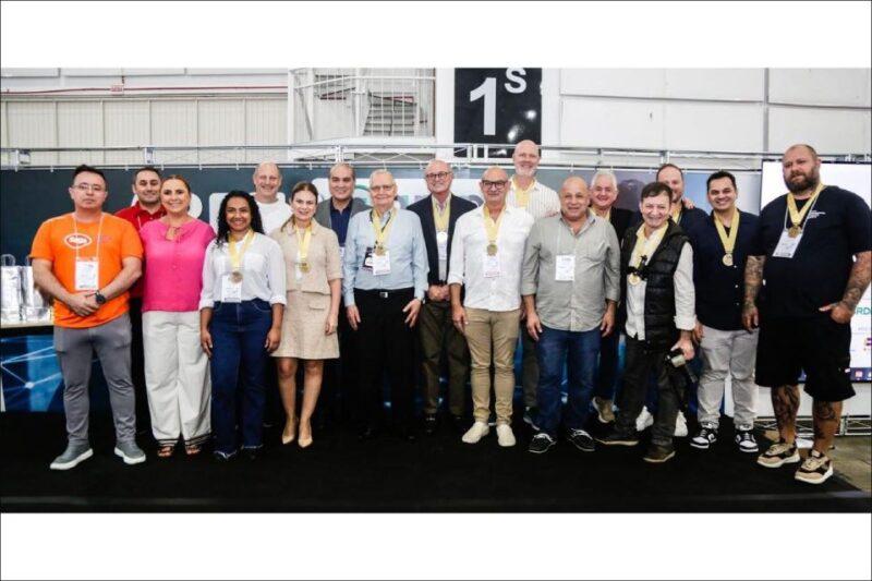 Foto de um grupo de 16 pessoas em pé, lado a lado, em um ambiente interno, possivelmente durante um evento ou conferência. Os indivíduos apresentam diversidade de idade e gênero, vestindo uma mistura de roupas formais e casuais. Várias pessoas usam medalhas em volta do pescoço, indicando reconhecimento ou participação em alguma atividade. O fundo mostra um espaço industrial ou de exposição, com estruturas metálicas e banners visíveis, reforçando o clima de evento.