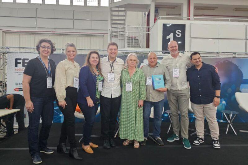 Grupo de oito pessoas sorrindo para a câmera durante um evento interno, posando em frente a um banner da Feira de Santa Catarina (FEAL). O local parece ser um espaço de exposições, com fundo de palco e algumas cadeiras. Uma das pessoas segura uma placa de reconhecimento, e todos usam crachás de identificação.