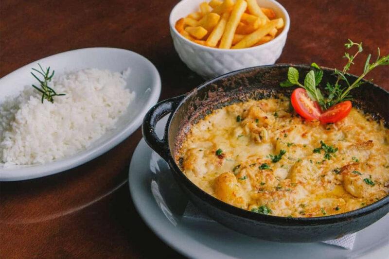 Prato de frutos do mar gratinado servido em uma panela de ferro com decoração de tomate cereja e ervas frescas. Acompanhado de uma tigela de arroz branco decorado com alecrim e uma tigela de batatas fritas. O conjunto está disposto sobre uma mesa de madeira.