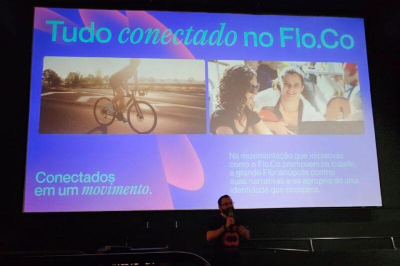 Apresentação do evento Floripa Conecta (Flo.Co), com um slide projetado em um telão. O slide apresenta o título 'Tudo conectado no Flo.Co' com imagens ilustrativas: à esquerda, uma pessoa andando de bicicleta ao pôr do sol; à direita, duas pessoas sorrindo em um ambiente descontraído. O texto na parte inferior do slide diz 'Conectados em um movimento. Na movimentação que iniciativas como o Flo.Co promovem na cidade, a Grande Florianópolis constrói suas narrativas e se apropria de uma identidade que prospera'. Em frente ao telão, uma pessoa fala ao microfone.