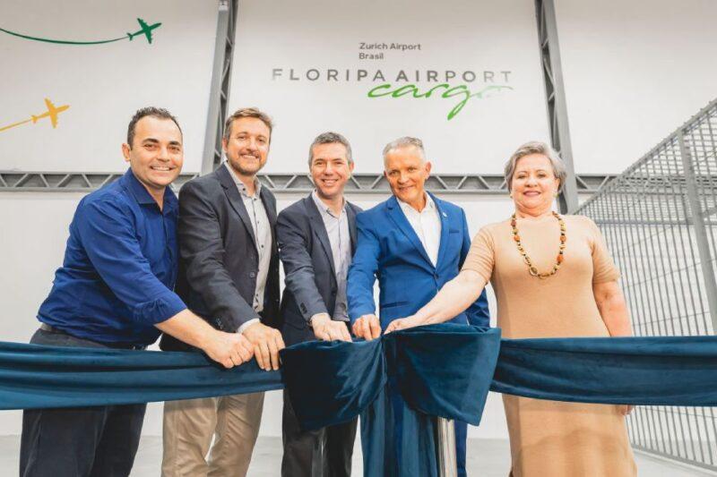 Cinco pessoas, quatro homens e uma mulher, participam de uma cerimônia de inauguração no Floripa Airport Cargo. Eles estão alinhados e segurando uma fita azul, prontos para cortá-la. Ao fundo, está o letreiro "Floripa Airport Cargo" e "Zurich Airport Brasil". A cena registra um momento formal e de celebração em um ambiente corporativo.