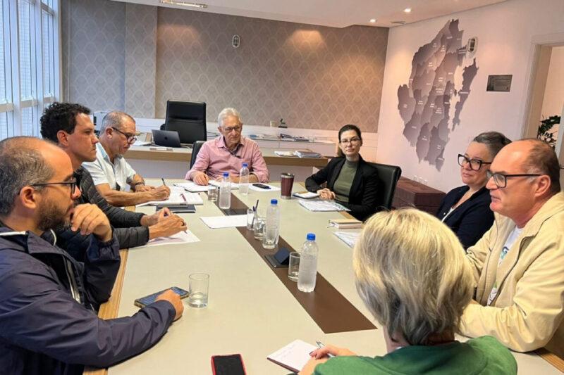 Reunião formal com oito pessoas sentadas em torno de uma mesa de conferência. A maioria dos participantes está atenta, com anotações e garrafas de água sobre a mesa. Ao fundo, um homem mais velho em uma cadeira de escritório observa a discussão. Na parede, há um grande mapa que destaca a região de Santa Catarina. O ambiente é de escritório, iluminado e profissional, sugerindo uma reunião de planejamento ou discussão estratégica.