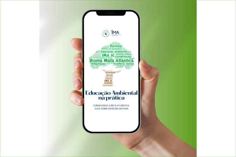 Uma mão segura um smartphone com a tela exibindo a capa de um guia digital intitulado 'Educação Ambiental na Prática'. O centro da imagem na tela do celular mostra uma árvore formada por palavras-chave relacionadas ao tema ambiental, como 'Bioma Mata Atlântica', 'restauração', 'IMA SC', 'florestas', 'conservação', 'espécies nativas', e outras. Acima da árvore está o logotipo do IMA (Instituto do Meio Ambiente de Santa Catarina). O fundo da imagem é gradiente em tons de verde, reforçando o tema ambiental.