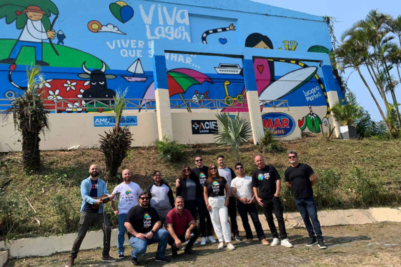 "Grupo de doze pessoas posando para uma foto em frente a um mural colorido em uma parede externa. O mural tem um tema alegre com desenhos de elementos como um pescador, um barco, um guarda-sol, e vários corações, acompanhados do texto 'Viva Lagoa' e 'Viver é mais que sonhar'. As pessoas na foto estão sorrindo e vestem roupas casuais, algumas com camisetas pretas e brancas com o logo 'Viva Lagoa'. Ao redor, há vegetação com palmeiras pequenas e grama, e ao fundo algumas árvores altas."