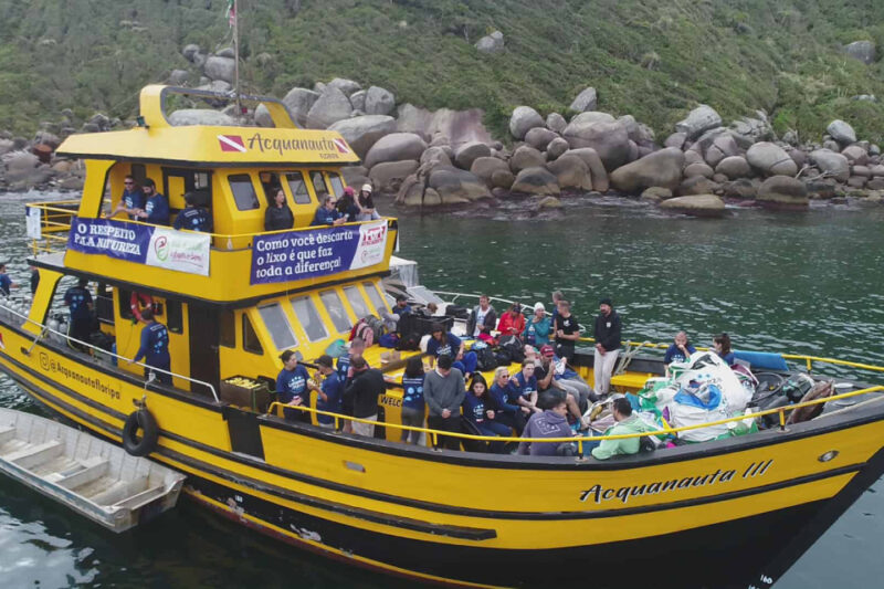 "Fotografia de um barco de cor amarela e preta, chamado 'Acquanauta III', ancorado em uma área de águas verdes e calmas próximas a uma costa rochosa. O barco está cheio de pessoas, muitas vestindo camisetas azuis que parecem estar participando de uma ação ambiental. Há várias sacolas grandes cheias de lixo coletado, provavelmente durante uma operação de limpeza dos mares. Duas faixas estão penduradas na lateral do barco, com as mensagens 'O respeito pela natureza' e 'Como você descarta o lixo é que faz toda a diferença!'. Ao fundo, a costa é composta por grandes rochas e vegetação verdejante."