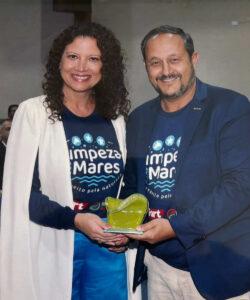 "Fotografia de um homem e uma mulher sorrindo, ambos segurando juntos um troféu de vidro verde em formato abstrato. A mulher, à esquerda, tem cabelo castanho cacheado e veste uma camiseta azul marinho com o logo 'Limpeza dos Mares' e uma jaqueta branca. O homem, à direita, tem cabelo curto e barba, e também veste uma camiseta azul marinho com o mesmo logo e um blazer azul escuro. Eles parecem estar em um evento de premiação ou celebração relacionada à proteção ambiental ou à limpeza dos mares."