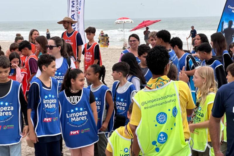 Um grupo de jovens voluntários participando de uma ação de limpeza de praia. Eles vestem coletes azuis e laranjas com a inscrição "Limpeza dos Mares", enquanto se organizam na areia. Ao fundo, o mar e algumas barracas de praia são visíveis. A atividade faz parte de um projeto de conscientização ambiental, com foco na preservação dos oceanos. O clima é de engajamento e colaboração, com todos focados na causa ambiental.