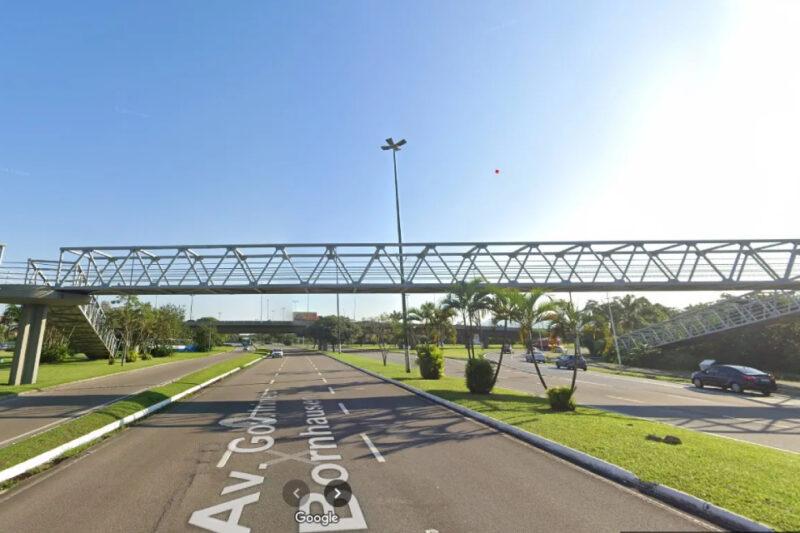 Passarela metálica para pedestres sobre a Avenida Governador Ivo Silveira, em Florianópolis, vista durante o dia sob um céu claro. A passarela é sustentada por pilares de concreto em ambos os lados, com rampas de acesso. Abaixo, a avenida de três faixas é ladeada por áreas verdes com pequenos arbustos e palmeiras. Poucos carros trafegam pela via, e um poste de iluminação está centralizado na imagem.