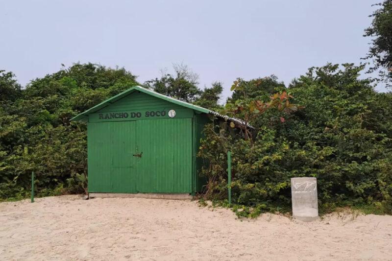 Uma pequena construção simples de madeira pintada de verde, com portas fechadas, está localizada em uma área de praia com areia clara. Acima das portas, está escrito "Rancho do Socó". A cabana é cercada por vegetação densa e árvores de diferentes tonalidades de verde, que quase a ocultam ao fundo. Um poste verde e um bloco de concreto estão posicionados à direita da cabana. O céu está nublado, criando um ambiente tranquilo e natural.