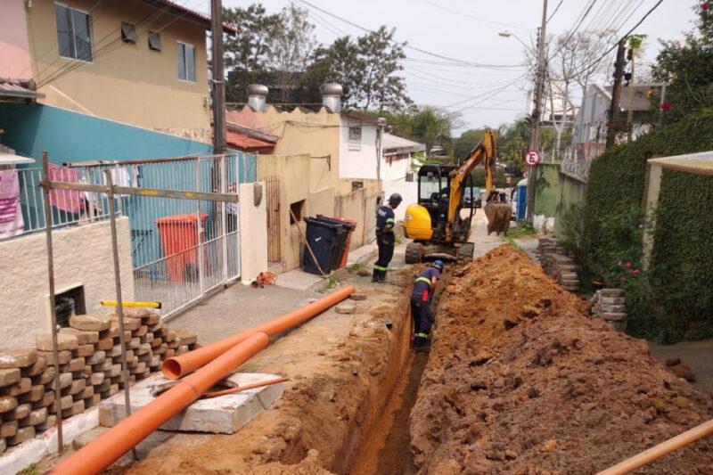 Trabalhadores de construção escavam uma vala em uma rua residencial estreita para a instalação de uma tubulação de esgoto laranja. Uma escavadeira amarela está sendo utilizada no processo, enquanto dois operários monitoram o trabalho. Ao redor da escavação, vê-se pilhas de terra, blocos de pavimentação e tubulações ainda por serem instaladas. As casas ao redor são de cores vibrantes, com muros pintados em tons de azul e amarelo. Redes elétricas e de telefonia são visíveis acima da rua, criando um cenário típico de bairro urbano.